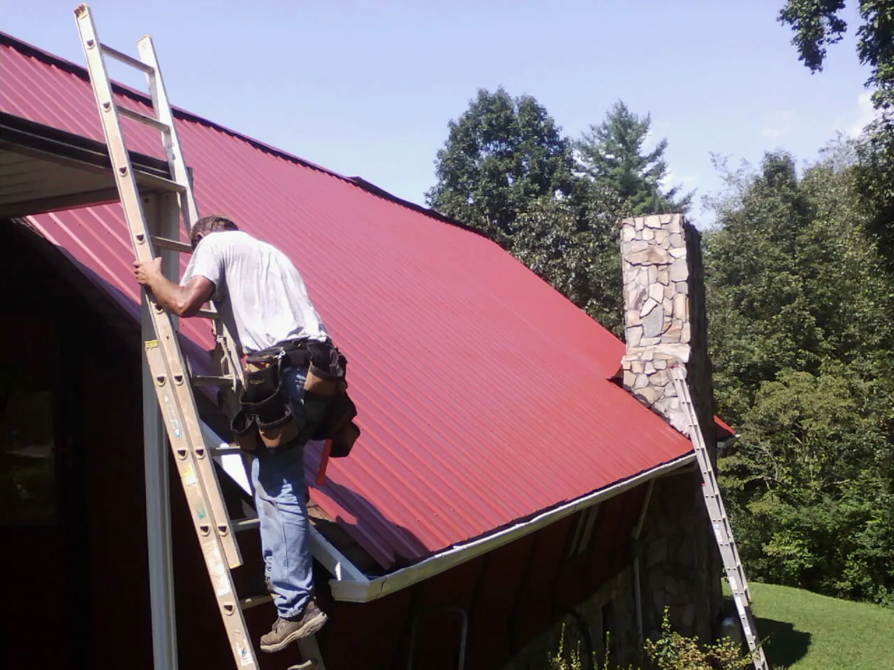 Roofer on a ladder beside a red metal roof and stone chimney — Soffit & Fascia Repair by Roofing Brothers in Frederick